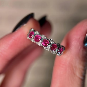 May include: A silver-toned band ring featuring a row of five round, vibrant pinkish-red gemstones set in a line. The band is accented with small, clear, sparkling stones nestled between and around the larger pinkish-red circular stones.