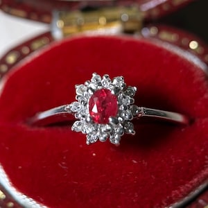 May include: A silver-toned ring featuring a central oval-cut red gemstone surrounded by a halo of small, clear, round-cut stones. The ring is set against a vibrant red velvet background, highlighting the contrast between the metal and the stones.