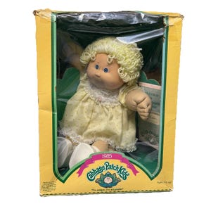 Puede incluir: Una muñeca Cabbage Patch Kids vintage en su caja original. La muñeca tiene cabello rubio rizado, ojos azules y viste un vestido amarillo con ribete de encaje blanco. La caja es amarilla y verde con el logo de Cabbage Patch Kids y el texto "1985".