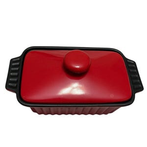 May include: A rectangular red ceramic baking dish with a matching lid and a black rim. The lid has a round knob. The dish has black handles on each end. The dish is designed for baking and serving.