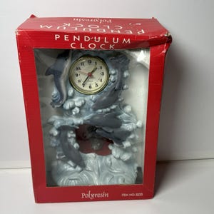 May include: A pendulum clock in a red box. The clock has a gold-coloured face with black numbers and hands. The clock is surrounded by a grey sculpture of dolphins leaping through waves. The box reads "PENDULUM CLOCK" and "Polyresin."