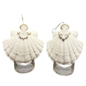 Puede incluir: Dos adornos de ángel de concha blanca. Cada ángel tiene un cuerpo de concha, una cabeza pequeña y un elemento decorativo en el pecho. Un ángel sostiene un copo de nieve y el otro una cruz. Ambos tienen colgadores de hilo dorado y se apoyan en soportes transparentes.