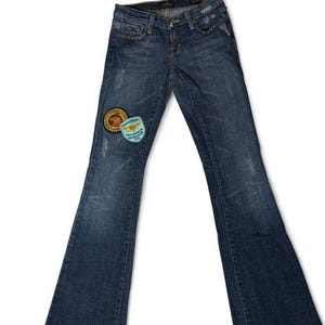 May include: Dark wash denim jeans with a flared leg. The jeans feature decorative rivets along the pockets and two patches on the left leg. One patch is round with an orange and yellow design, and the other is a blue and white patch with text.
