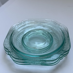 May include: A stack of four clear, light blue glass dishes. The dishes have a square shape with rounded corners and a decorative floral pattern around the edges. The dishes are nested, with a smaller, circular dish at the center.