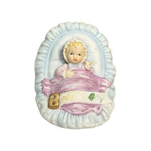 May include: A ceramic figurine of a baby in a light blue bassinet. The baby is wearing a pink outfit and a bonnet. A pink blanket covers the baby. The word "Baby" is written on a banner across the blanket.