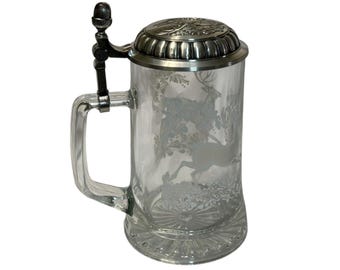 May include: Clear glass beer stein with a metal lid and handle. The glass is etched with a forest scene featuring deer. The lid has a decorative design. The stein has a large handle and a detailed base.