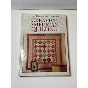May include: A book titled "Creative American Quilting" by Better Homes and Gardens. The cover features a detailed quilt design with various colors and patterns. A small framed picture and a woven basket are also visible.