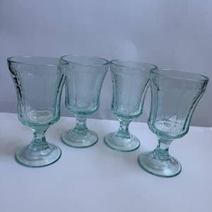 May include: Four clear, stemmed glasses with a light blue tint. Each glass has a textured, leaf-like pattern and a rounded bowl. The glasses are arranged on a white surface, showcasing their elegant design.