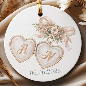May include: A round white ceramic ornament featuring a gold-toned heart-shaped locket illustration with the letters A and N inside. Above the locket is a soft pink ribbon with floral accents. The date 06.06.2026 is printed in gray below the locket.