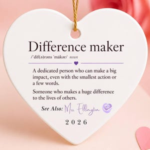 May include: A white heart-shaped ceramic ornament hanging by a gold cord. It features the text Difference maker, a definition of the term, the name Mrs. Ellington in script, a small purple rose icon, and the year 2026 printed in black ink.