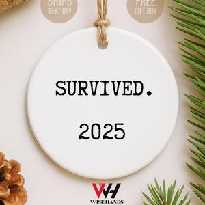 May include: A white ceramic ornament with the text "SURVIVED. 2025" in black. The ornament is round and has a string for hanging. The Wise Hands logo is at the bottom. The background includes pine needles and a pine cone.