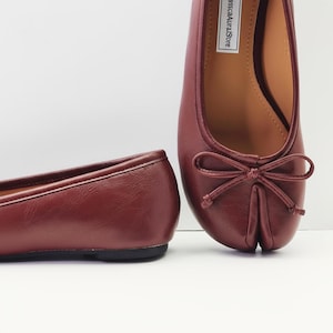 May include: Two burgundy ballet flats are shown, with a rounded toe and a small bow detail. The shoes are made of a smooth material and have a low heel. The background is white. The shoes are a deep red colour.