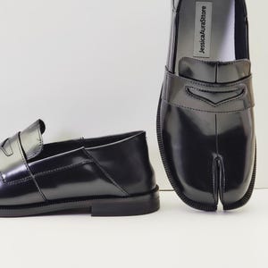 Pode incluir: Mocassins pretos de couro com bico dividido. Os sapatos têm um acabamento brilhante e um design clássico de mocassim com borlas, com um detalhe único de bico dividido. O nome da marca "JessicaAuraSttore" está impresso na palmilha.