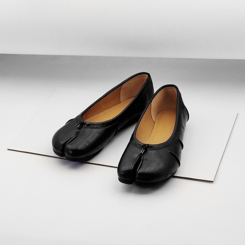 May include: A pair of black leather split-toe ballet flats. The shoes have a zip detail on the front and a tan interior. The shoes are displayed on a white surface.