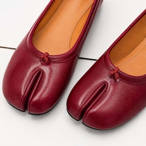 May include: A pair of burgundy leather split-toe ballet flats. The shoes have a unique design with a separate compartment for the big toe. The interior is tan, and a small knot detail adorns the top.