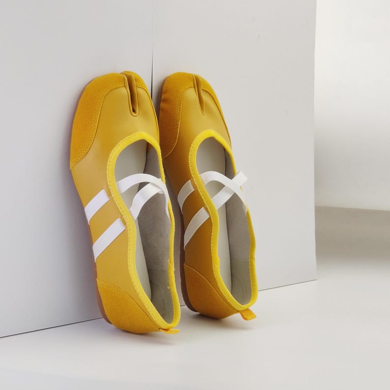 Puede incluir: Un par de zapatos amarillos con puntera dividida, rayas y correas blancas. Los zapatos tienen un tal&oacute;n y una puntera de ante. El dise&ntilde;o recuerda al calzado deportivo.