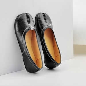 Op de afbeelding: Een paar zwarte leren split-teen flats met witte stiksels en een kleine decoratieve bloem. De schoenen hebben een gele binnenzool en een ronde neus. De flats leunen tegen een witte muur.