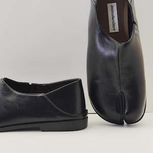 May include: Black leather split-toe shoes. The shoes have a low heel and a unique design with a split between the big toe and the other toes. The shoes are displayed on a white surface. The text "JessicaAuraStore" is visible on the inner sole.