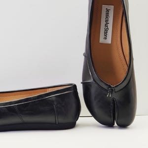 May include: Black leather ballet flats with a split toe design. The shoes have a rounded toe and a low heel. The brand name "JessicaAurStore" is printed on the insole.