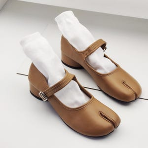 May include: A pair of brown leather Tabi shoes with a strap and buckle. The shoes have a split toe design and a low heel. White socks are inserted into the shoes.
