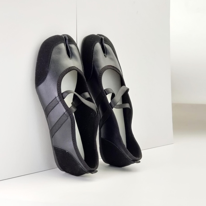 Puede incluir: Un par de zapatos negros con puntera dividida, con una correa y una separaci&oacute;n para el dedo gordo. Los zapatos combinan materiales lisos y de tipo ante, mostrando un dise&ntilde;o minimalista. Miden aproximadamente 25 cm de largo.
