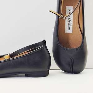 Può includere: Ballerine in pelle nera con design a punta divisa e cinturino alla caviglia. Le scarpe hanno un tacco basso e una chiusura con fibbia. La soletta è di colore marrone chiaro e il marchio "Jessica AirraStore" è visibile.