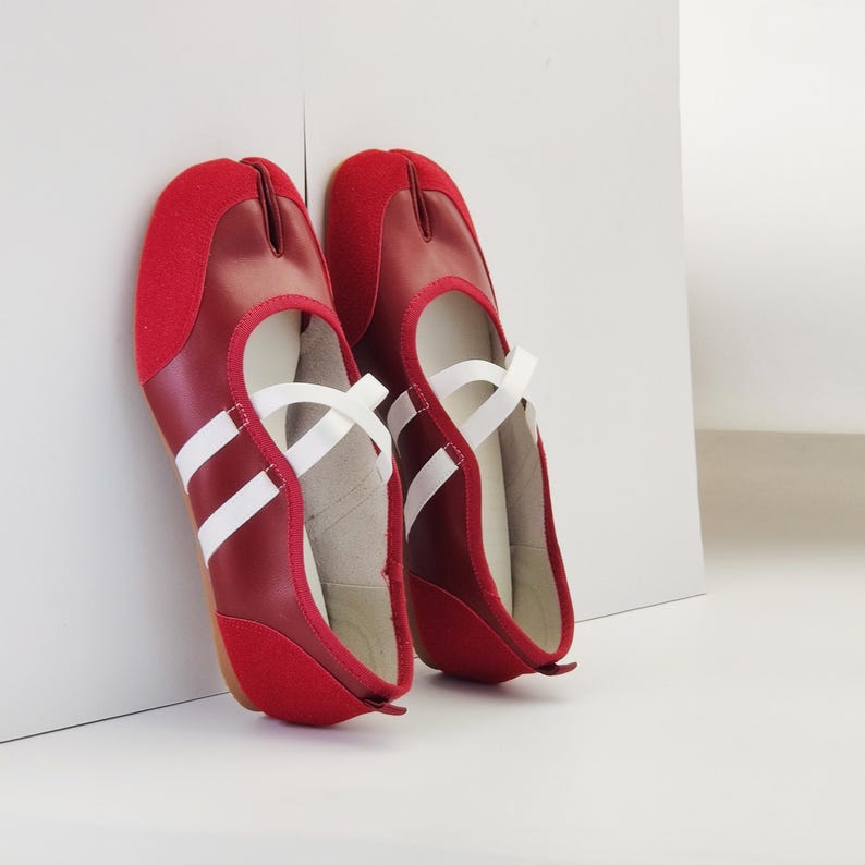 Puede incluir: Un par de zapatos rojos con puntera dividida y detalles blancos. Los zapatos tienen un dise&ntilde;o de correas y una raya blanca en contraste. Los zapatos est&aacute;n apoyados contra una pared blanca.