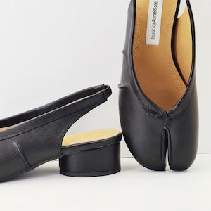 Op de afbeelding: Zwarte leren slingback schoenen met een split-teen ontwerp en een kleine blokhak. De schoenen hebben een minimalistische stijl en een neutraal kleurenpalet, geschikt voor diverse outfits. De binnenzool is bruin.