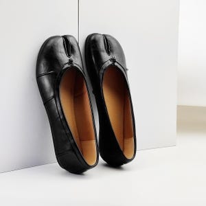 May include: A pair of black split-toe ballet flats. The shoes are made of smooth leather and feature a split between the big toe and the other toes. The interior is a light brown color. The shoes are leaning against a white wall.