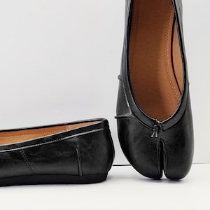 May include: Black leather split-toe ballet flats. The shoes have a rounded toe and a split design for the big toe. The interior is a light brown color. The shoes are on a white background.