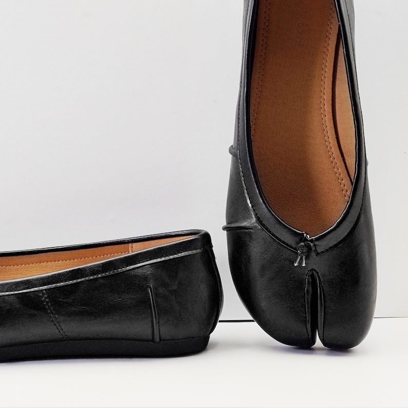May include: Black leather split-toe ballet flats. The shoes have a rounded toe and a split design for the big toe. The interior is a light brown color. The shoes are on a white background.