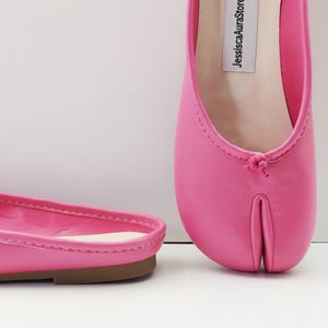 May include: Bright pink split-toe ballet flats. The shoes have a rounded toe design with a small knot detail at the top. The sole is a light brown color. The brand name "JessicaAuraStore" is printed on the insole.