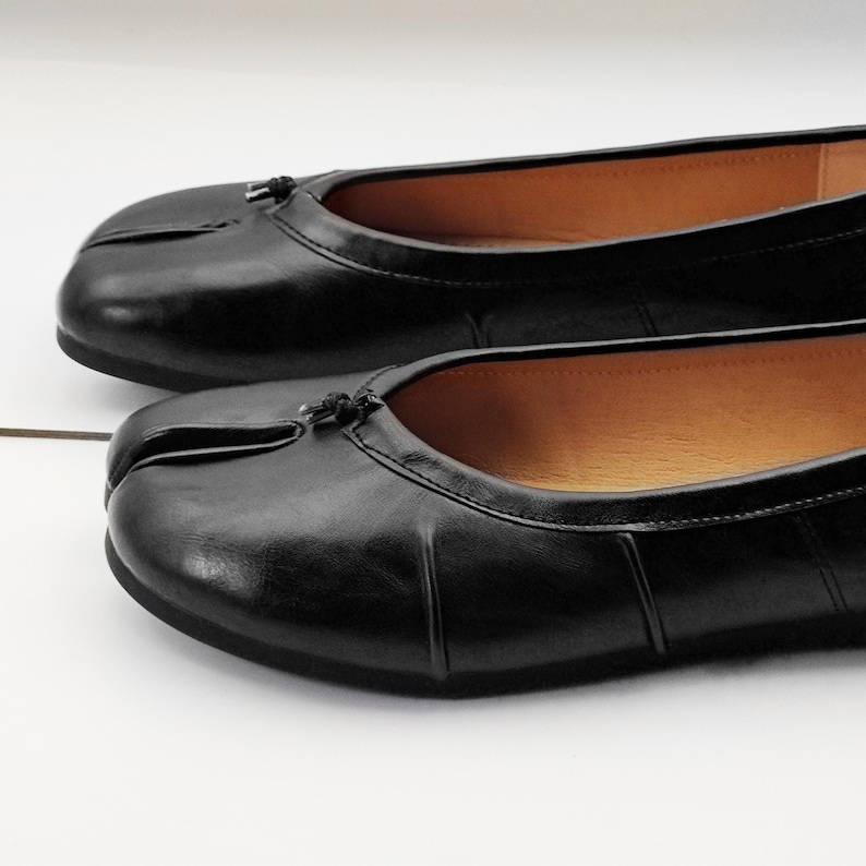 May include: A pair of black leather ballet flats. The shoes have a split-toe design and a small bow detail at the top. The interior of the shoe is a light brown color. The shoes are on a white surface.