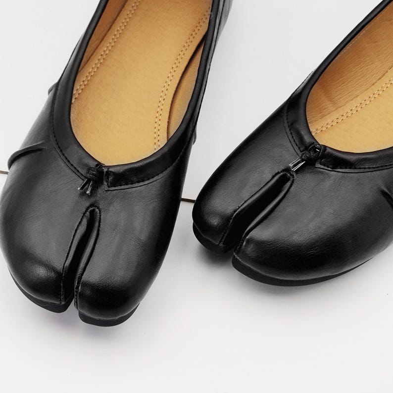 May include: Black leather split-toe ballet flats. These flats feature a unique design with a split between the big toe and the other toes. The shoes have a rounded toe and a small bow detail. The interior is a light tan color.