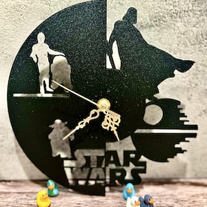 May include: Black Star Wars themed wall clock with a circular design. The clock features silhouettes of characters and the Death Star. The words "STAR WARS" are at the bottom. Gold clock hands and a gold center piece.
