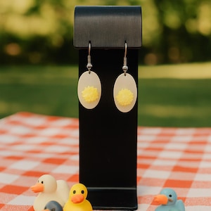 May include: Oval-shaped earrings with a yellow flower design are displayed on a black stand. The earrings are showcased with a collection of small, colorful rubber duck figurines on a red and white checkered picnic blanket.