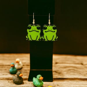 May include: A pair of frog-shaped earrings with a bright green body and black outlines. The earrings are hanging from gold-colored hooks. Small, colorful duck figurines are in the foreground.