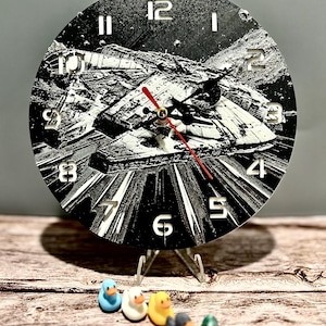 May include: A black and white round clock with a spaceship design, featuring a red second hand. The clock is displayed on a clear stand, with a row of colorful rubber ducks in front. The clock's diameter is approximately 8 inches.