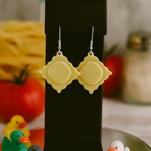 May include: A pair of yellow ravioli-shaped earrings with silver hooks are displayed on a black stand. The earrings have a square shape with a circular center. The background includes a tomato, pasta, and toy ducks.