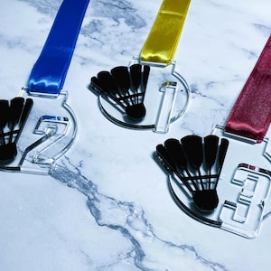 May include: Clear acrylic medals with black badminton shuttlecock designs and colored ribbons. The medals are numbered 1, 2, and 3, with blue, yellow, and burgundy ribbons, respectively. The medals are on a marble surface.