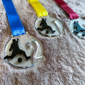 May include: Three clear acrylic soccer medals with colorful ribbons. Each medal features a black silhouette of a soccer player kicking a ball. The ribbons are blue, yellow, and pink, adding a vibrant touch to the awards.