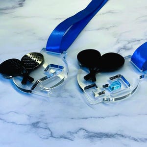 May include: Two clear acrylic medals with blue ribbons. Each medal features a black ping pong paddle design. One medal has a silver paddle with holes. The medals are on a marble surface.