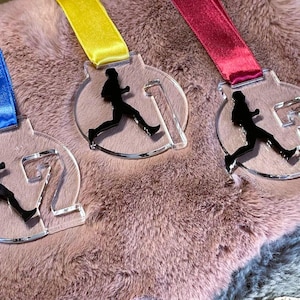 May include: Three clear acrylic medals with black runner silhouettes and numbers 1, 2, and 3. Each medal has a different colored ribbon: blue, yellow, and red. The medals are displayed on a pink, fluffy surface.