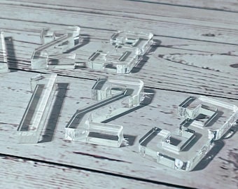 May include: Clear acrylic numbers 1, 2, and 3 are scattered on a light wood-grain surface. The numbers are blocky and three-dimensional, casting shadows. The numbers are transparent, allowing the wood grain to be seen through them. The numbers are likely for crafts or decor.