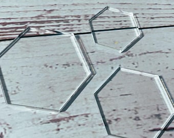 May include: Three clear, geometric, hexagonal acrylic shapes of varying sizes. The shapes are transparent, allowing light to pass through, and are arranged on a light-colored, wooden surface. The edges of the shapes are sharply defined.