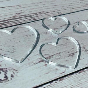 Könnte beinhalten: Vier klare Acrylherzformen in verschiedenen Größen liegen auf einer hellen Holzoberfläche. Die transparenten Herzen lassen die Holzmaserung durchscheinen. Diese Herzen sind wahrscheinlich für Bastel- oder Dekorationszwecke gedacht.