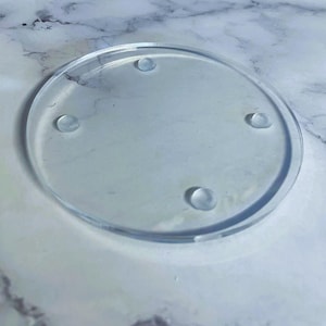 Può includere: Un sottobicchiere rotondo e trasparente con quattro piccoli piedini arrotondati. Il sottobicchiere è progettato per proteggere le superfici dalla condensa delle bevande. È mostrato su una superficie a motivo marmoreo. Diametro: circa 10 cm.