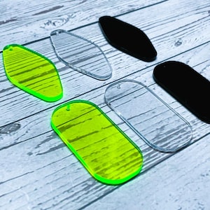 Op de afbeelding: Een assortiment acryl blanks in verschillende vormen en kleuren. De afbeelding toont twee heldere, twee zwarte en twee neon groene blanks. De groene blanks hebben een gestreept patroon. Elke blank heeft een klein gaatje om op te hangen.