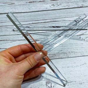 Puede incluir: Tiras rectangulares de acrílico transparente, algunas sujetas en la mano, otras dispersas sobre una superficie de madera clara. Las tiras son reflectantes, con un aspecto espejado. Los bordes son limpios y afilados, sugiriendo un diseño moderno.