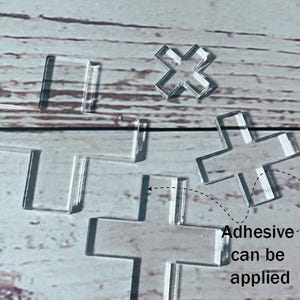 Könnte beinhalten: Klare, kreuzförmige Verzierungen aus Acryl. Das Bild zeigt mehrere Kreuze unterschiedlicher Größe, mit dem Text "Adhesive can be applied", der auf ihre mögliche Verwendung hinweist. Die Kreuze befinden sich auf einer hellen Holzmaserung.